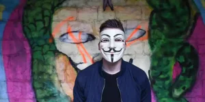 Person wearing an Anonymous-style Guy Fawkes mask standing in front of colorful graffiti, symbolizing anonymous reviews.